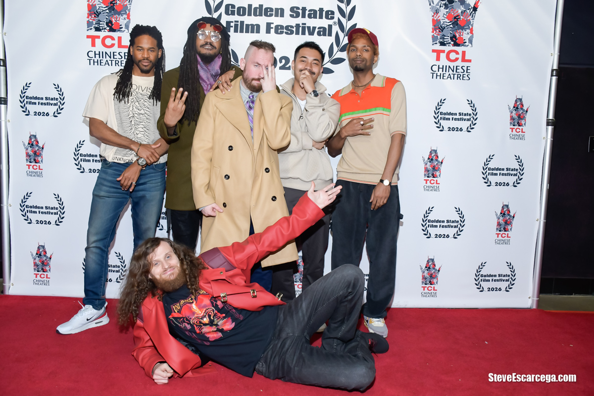 Group of six people posing on red carpet in front of a backdrop with festival logos, one person lying down in front.