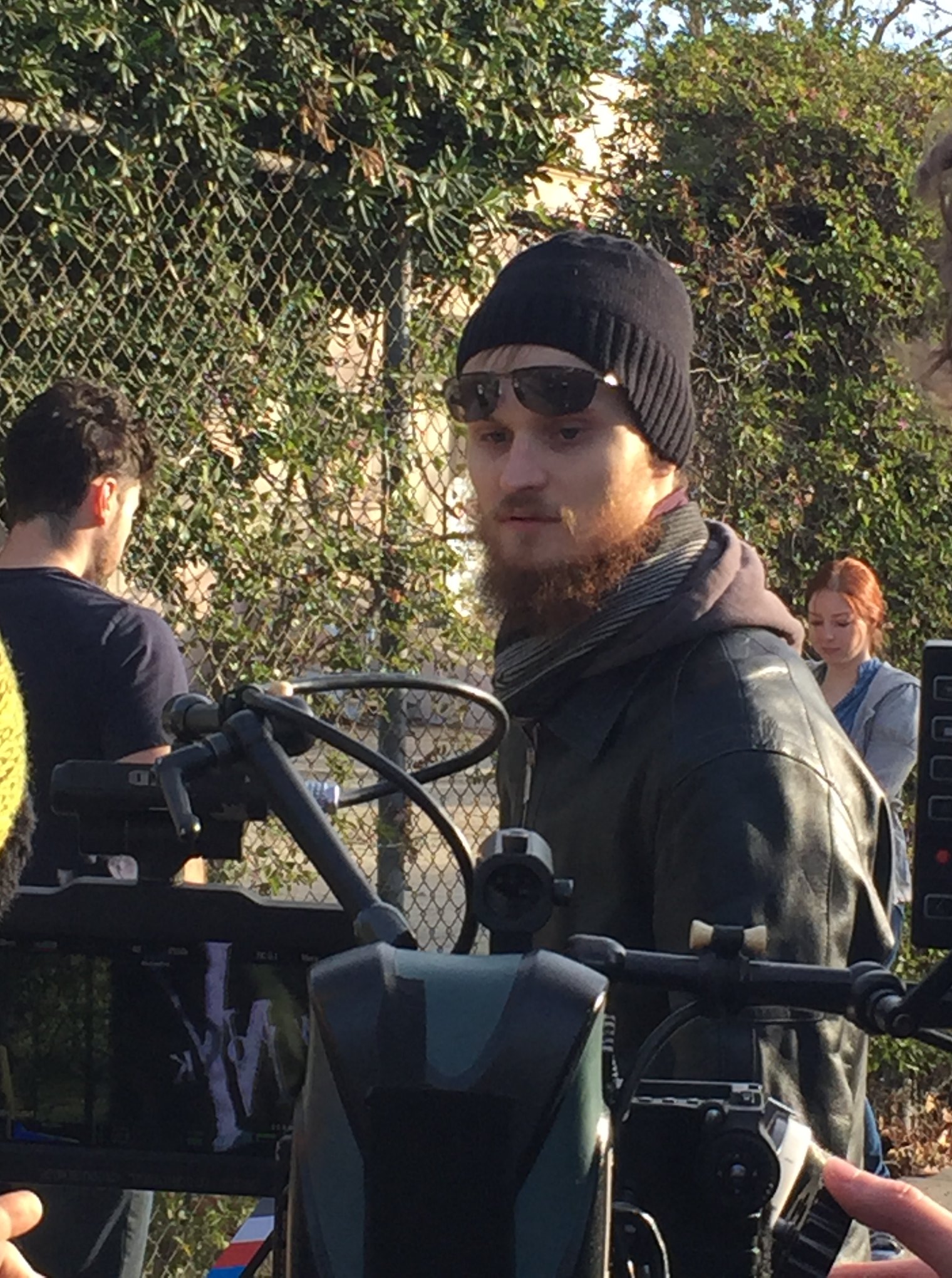 Young man with beard wearing sunglasses, black beanie, and leather jacket outdoors, with people and greenery in background.