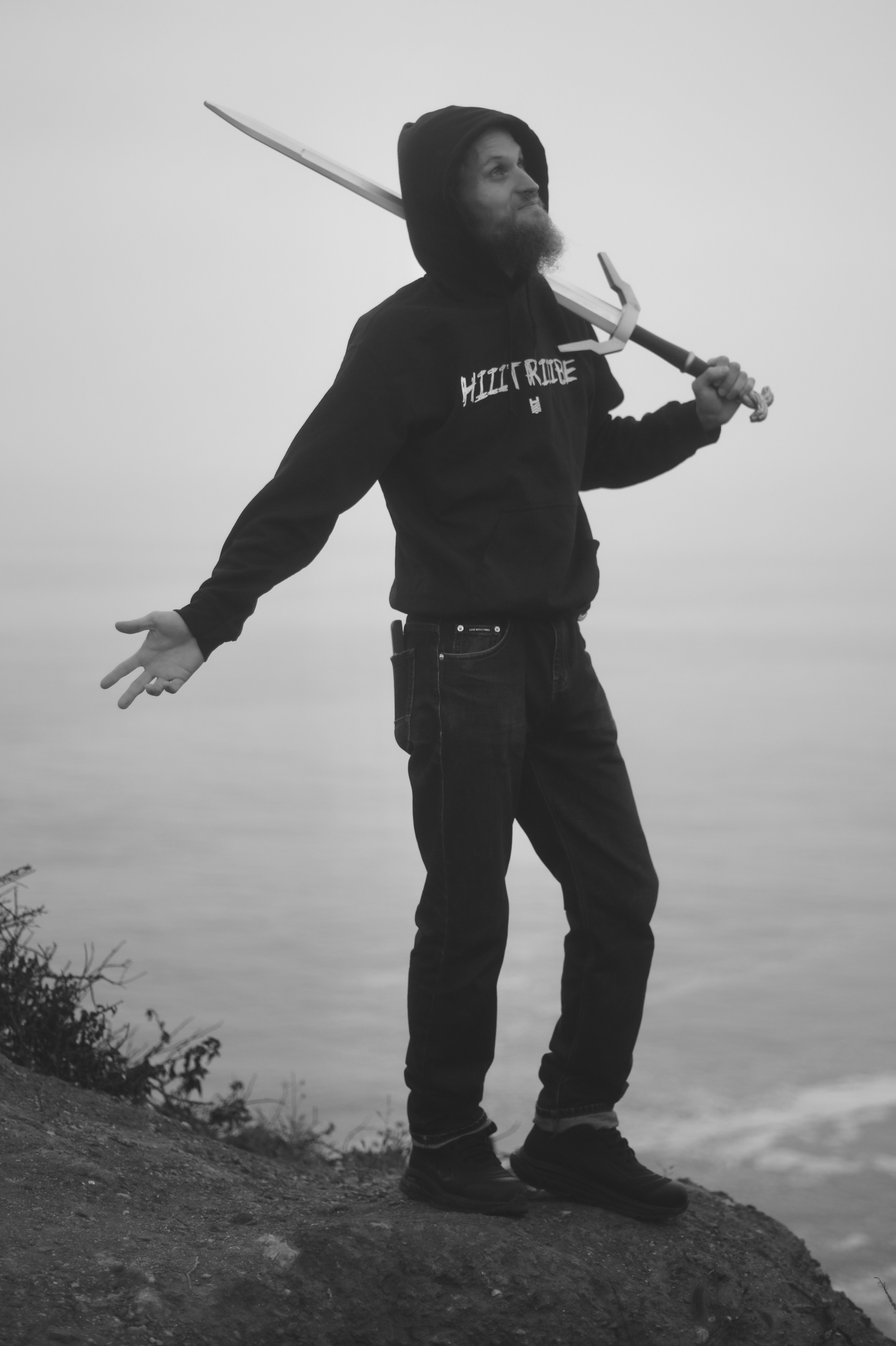 Person standing on a hill holding a sword over shoulder, arms outstretched, with a cloudy sky background.