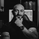 Man with a beard and tattooed arm, sitting with hand on chin, in a dark room with computer screens behind him.