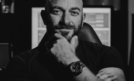Man with a beard and tattooed arm, sitting with hand on chin, in a dark room with computer screens behind him.