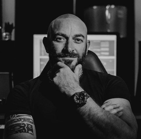 Man with a beard and tattooed arm, sitting with hand on chin, in a dark room with computer screens behind him.