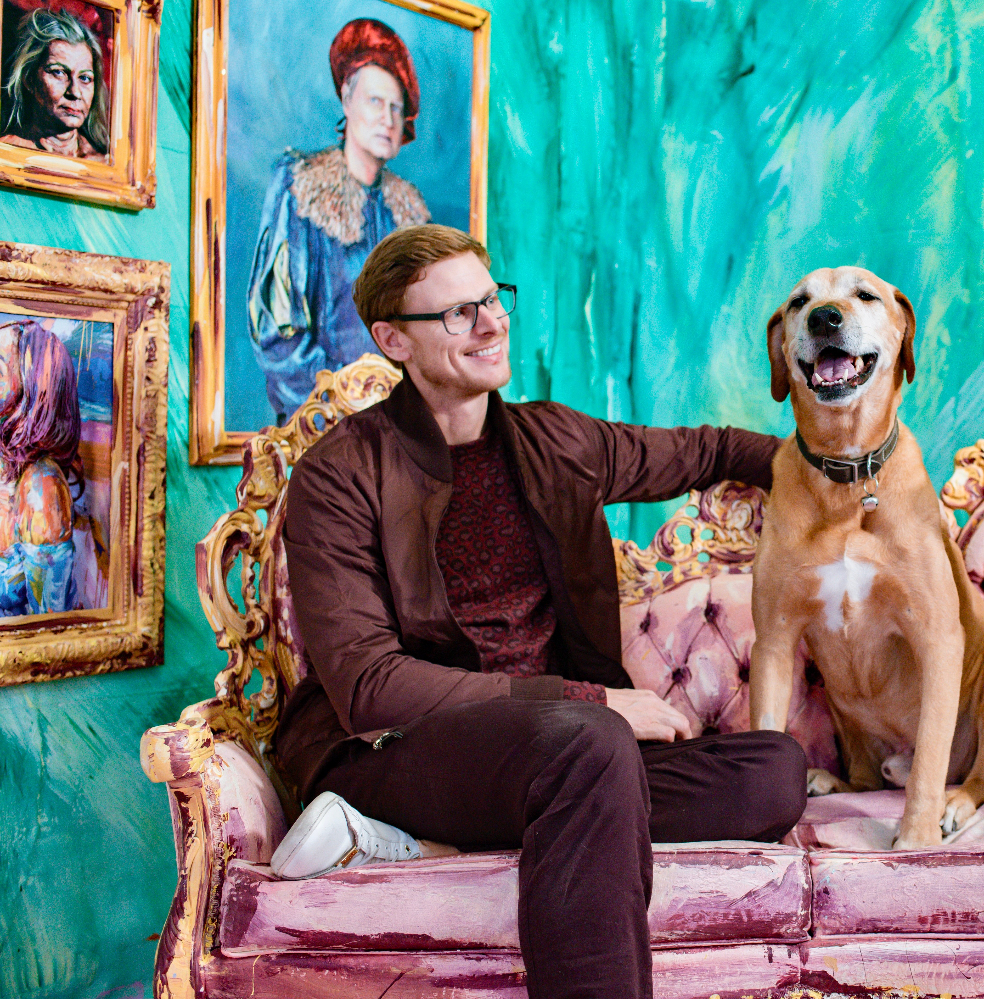 Man with glasses sitting on a pink vintage sofa with a large dog, colorful paintings on teal wall behind them.