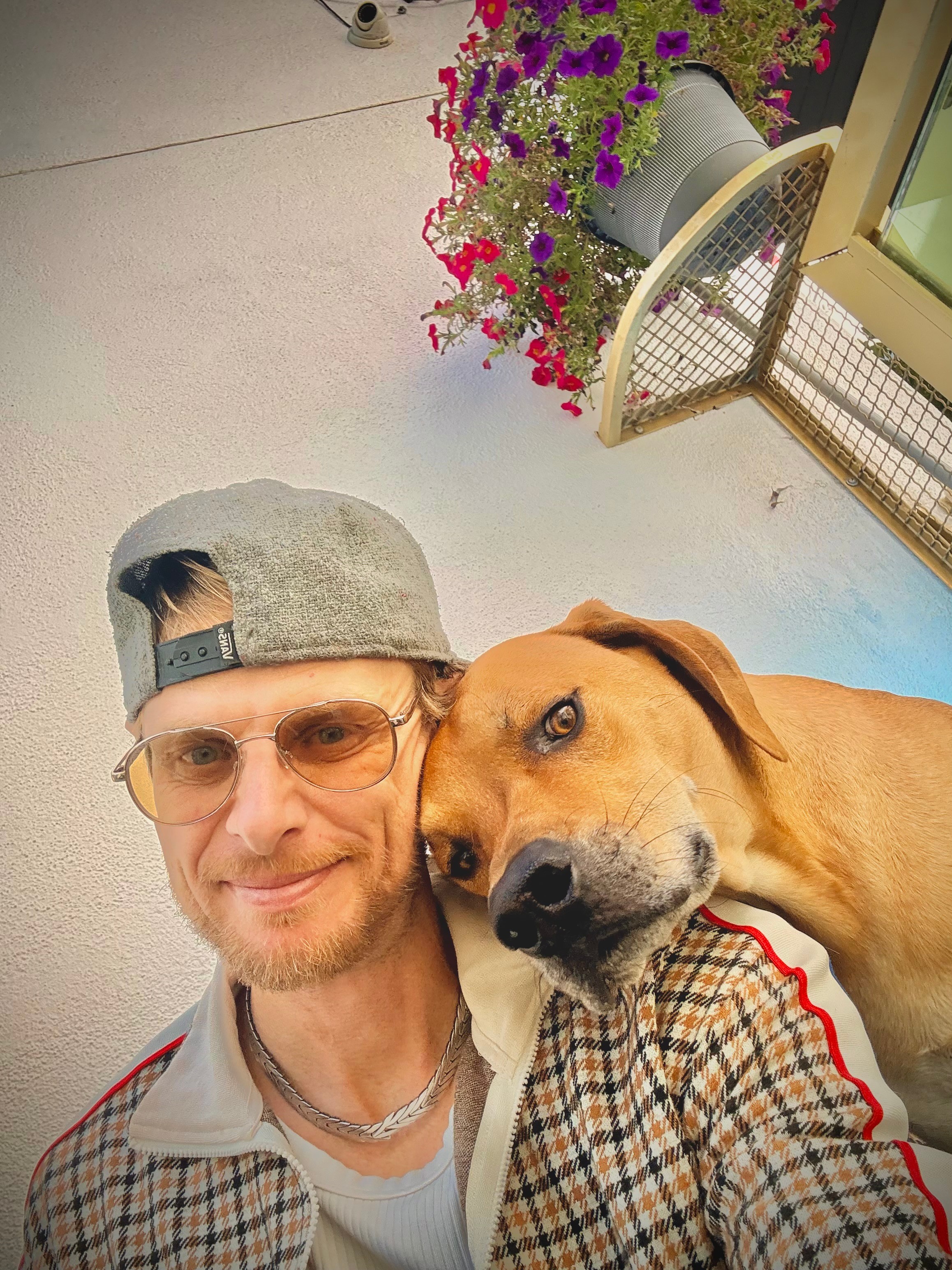 Person with glasses and a cap taking a selfie with a dog, outdoors near a flower pot and a window.