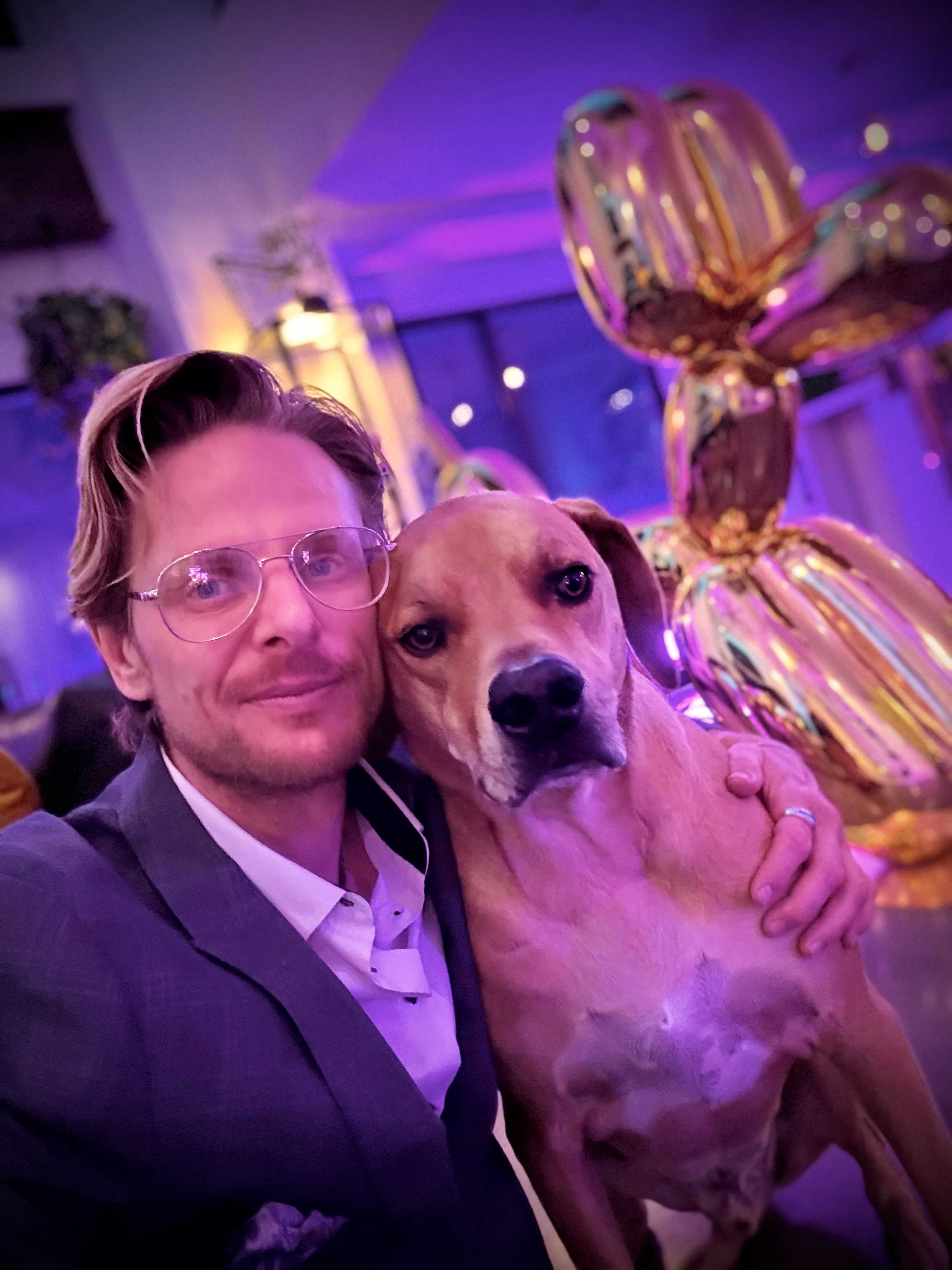 Man with glasses and a dog at an indoor event with colorful lighting and balloon decorations.
