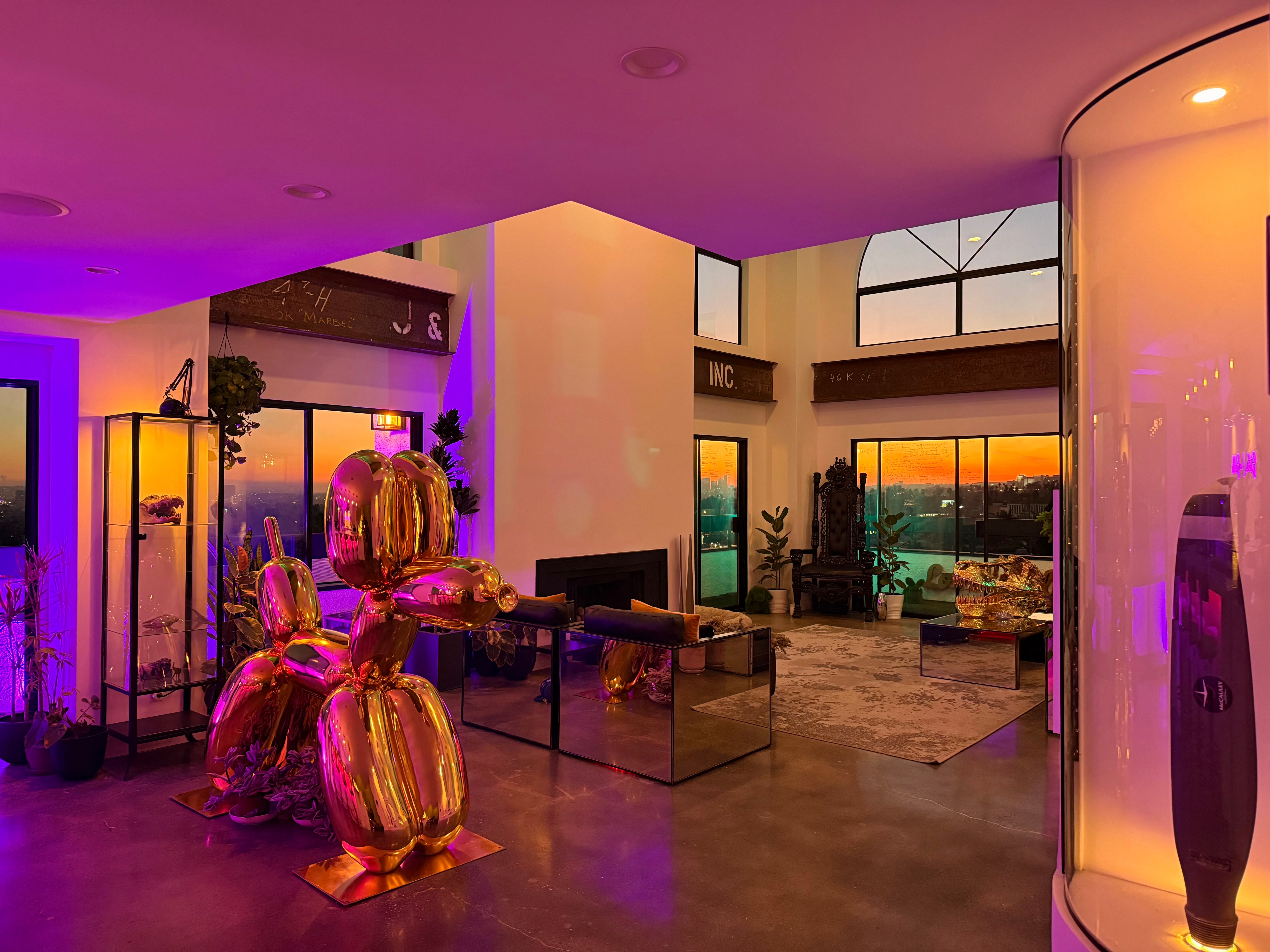 Interior of a modern room with colorful lighting, large windows showing a sunset, and decorative objects including vases and plants.