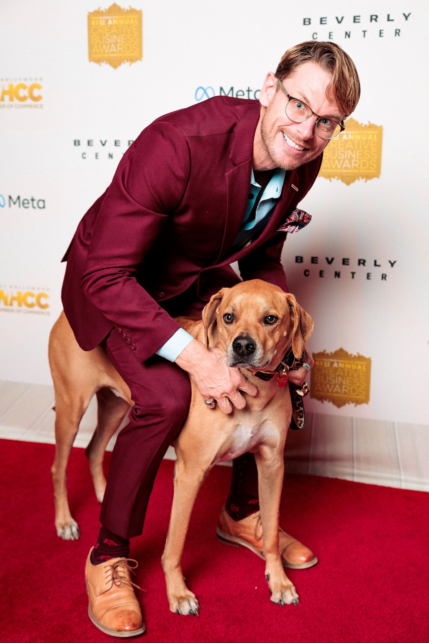 Man in maroon suit crouching next to a large brown dog on a red carpet, smiling, with a white backdrop featuring logos.