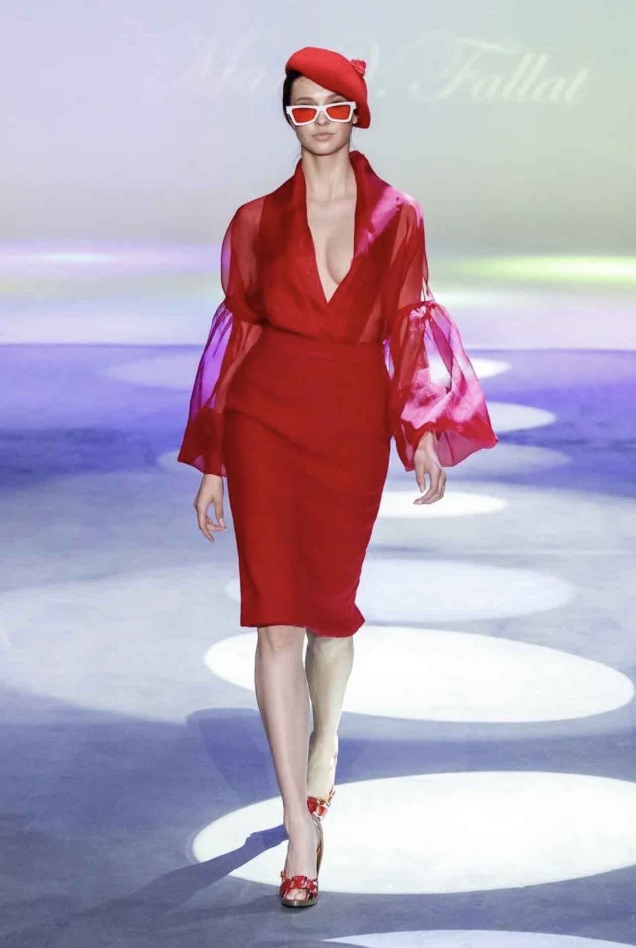 Model walking on runway wearing red dress, hat, and sunglasses, with a light background and runway lights.