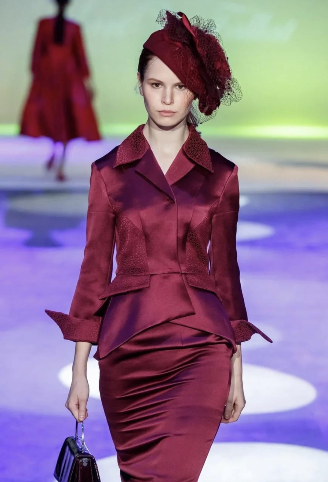 Model walking on runway wearing a burgundy outfit with a matching hat, holding a small handbag, background blurred.