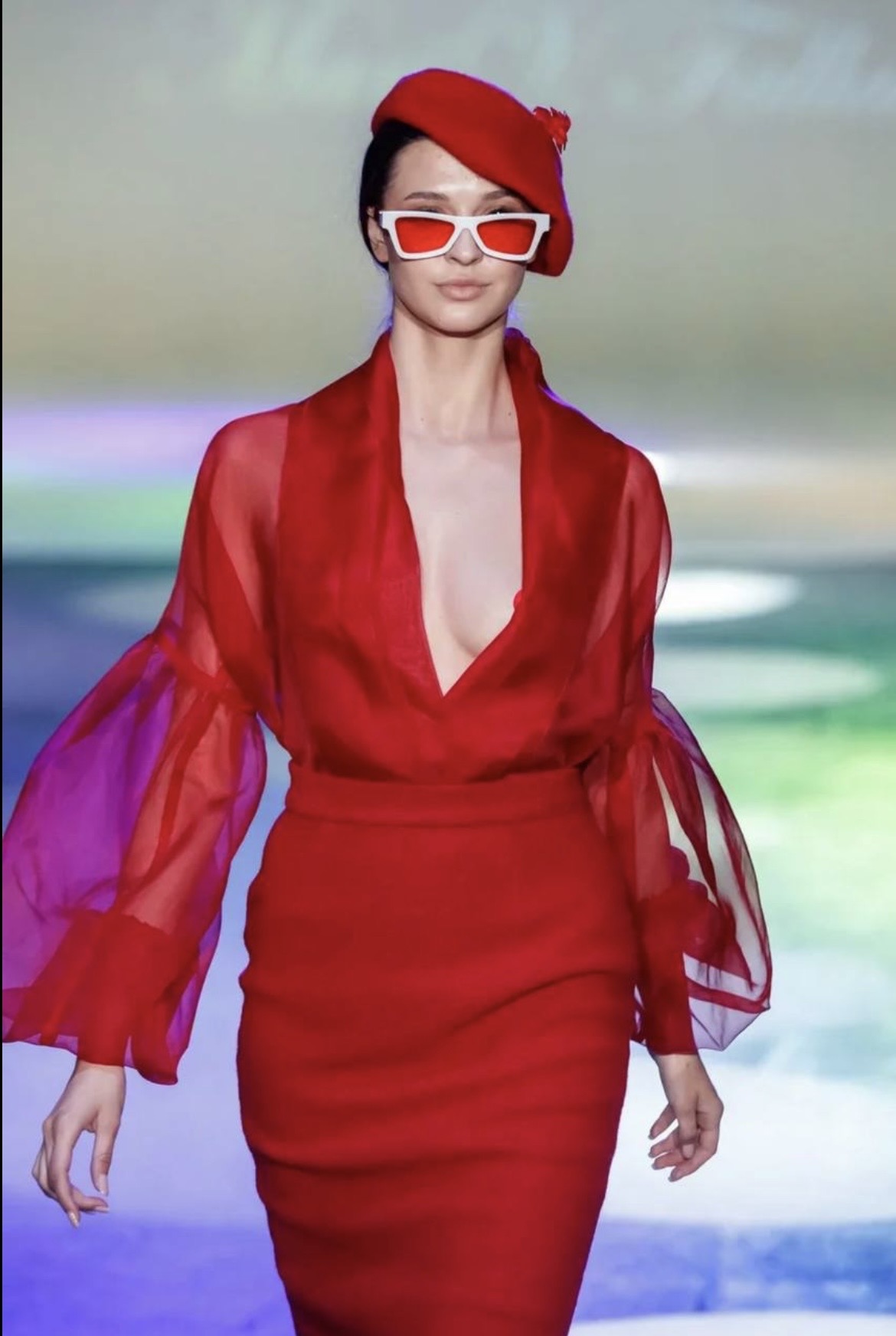 Woman in red dress with sheer sleeves, red hat, and white sunglasses walking on runway.
