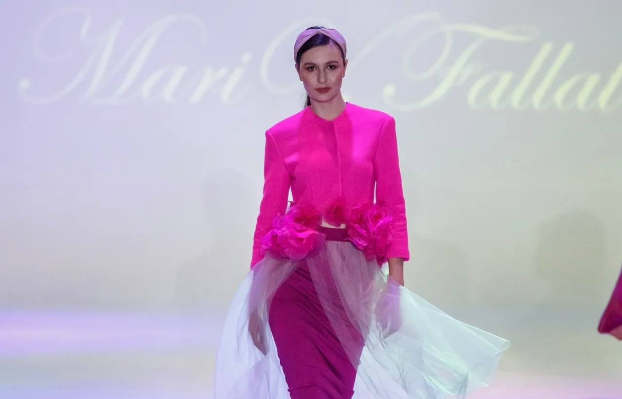 Model walking on runway wearing a bright pink jacket and a white tulle skirt, with a headband, against a light background.
