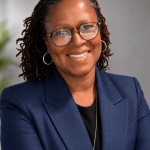 Portrait of a smiling woman with glasses, wearing a navy blazer and black top, against a neutral background.