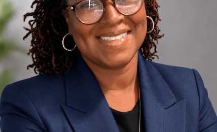 Portrait of a smiling woman with glasses, wearing a navy blazer and black top, against a neutral background.