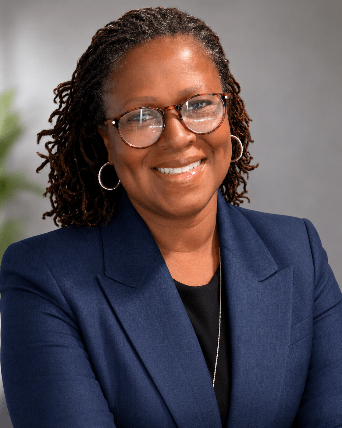 Portrait of a smiling woman with glasses, wearing a navy blazer and black top, against a neutral background.