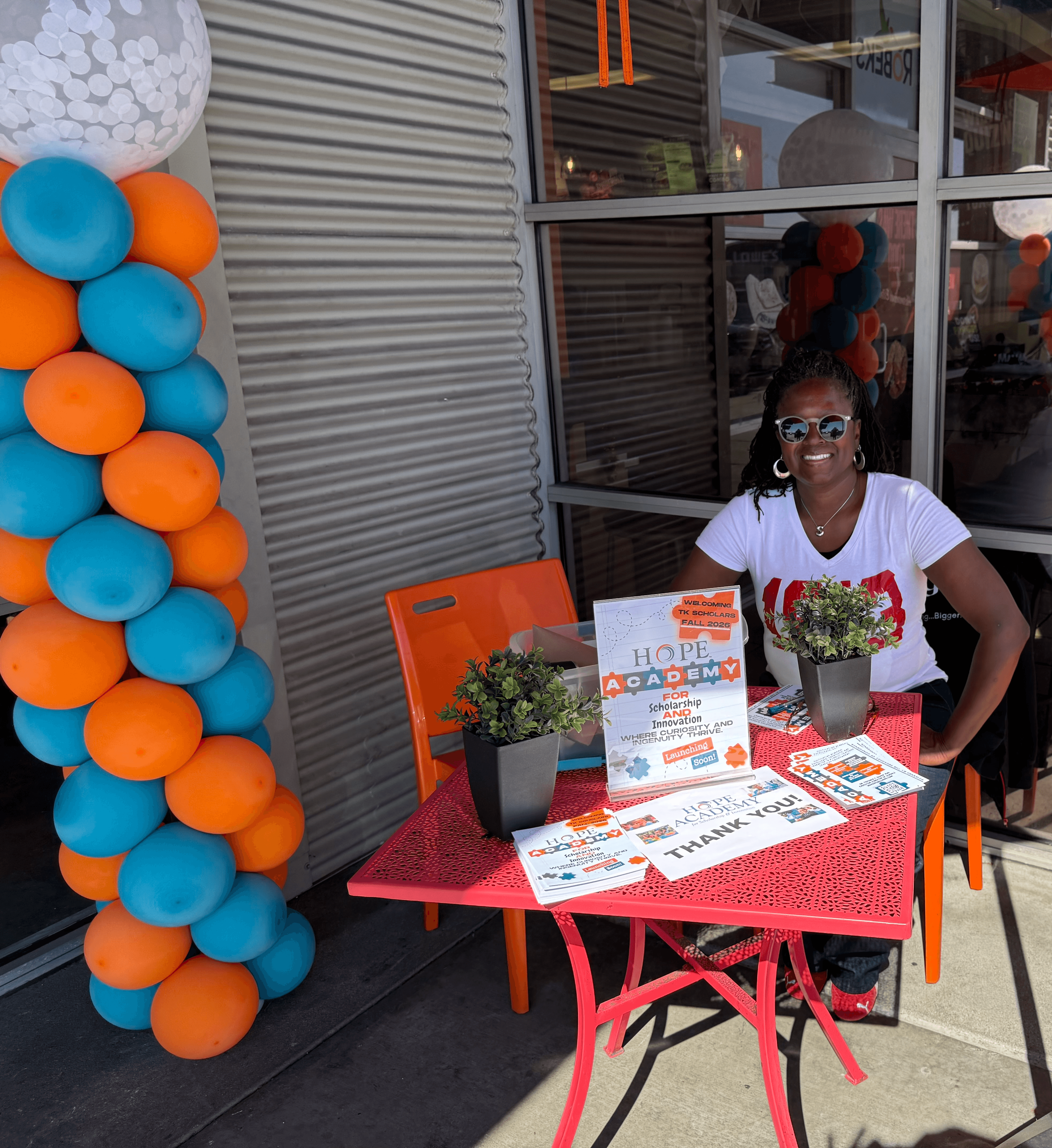 Smiling woman sitting at a red table with flyers and potted plants outside a building.