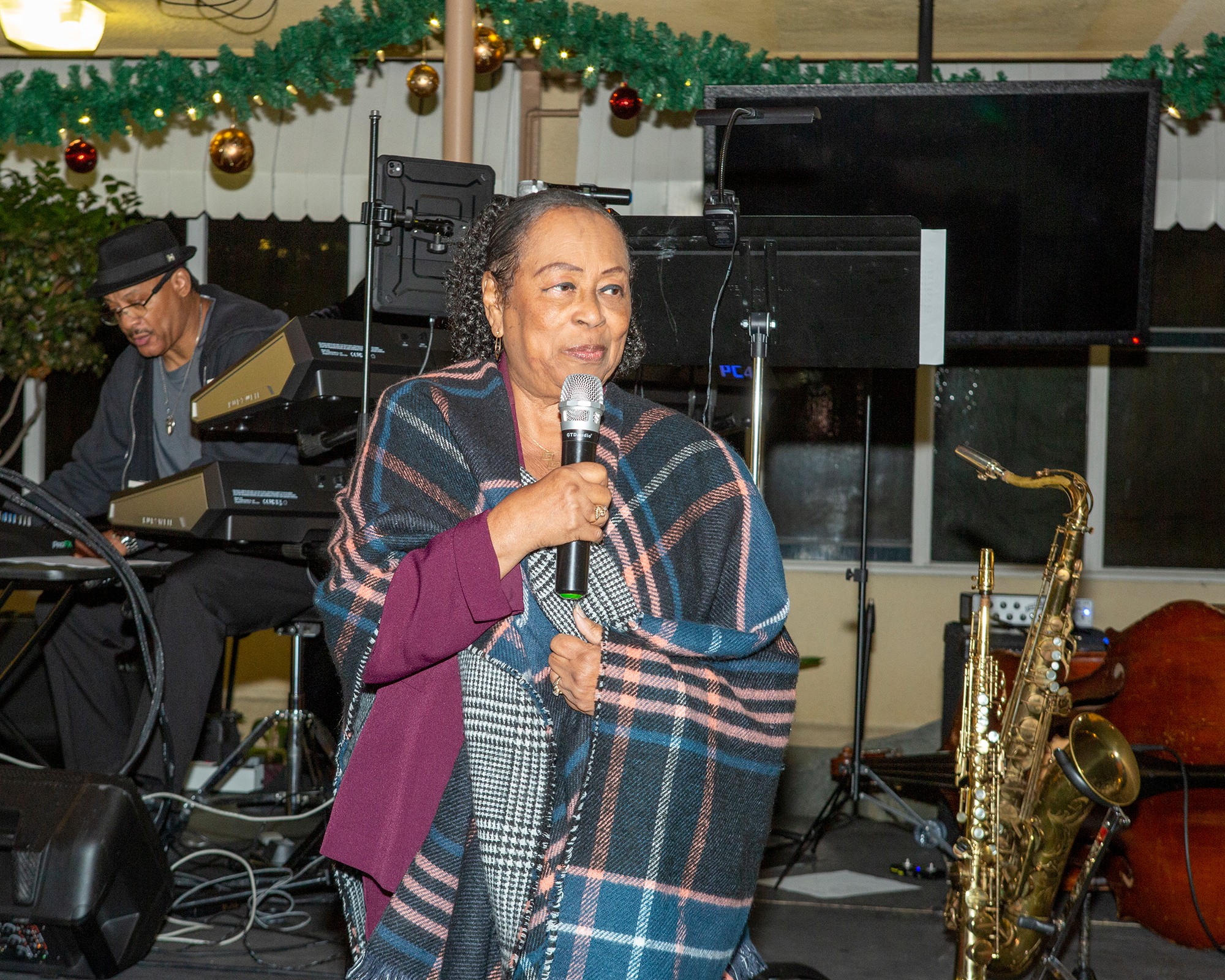 Woman holding microphone, standing in front of musical instruments and a man playing keyboard, decorated with Christmas garland.