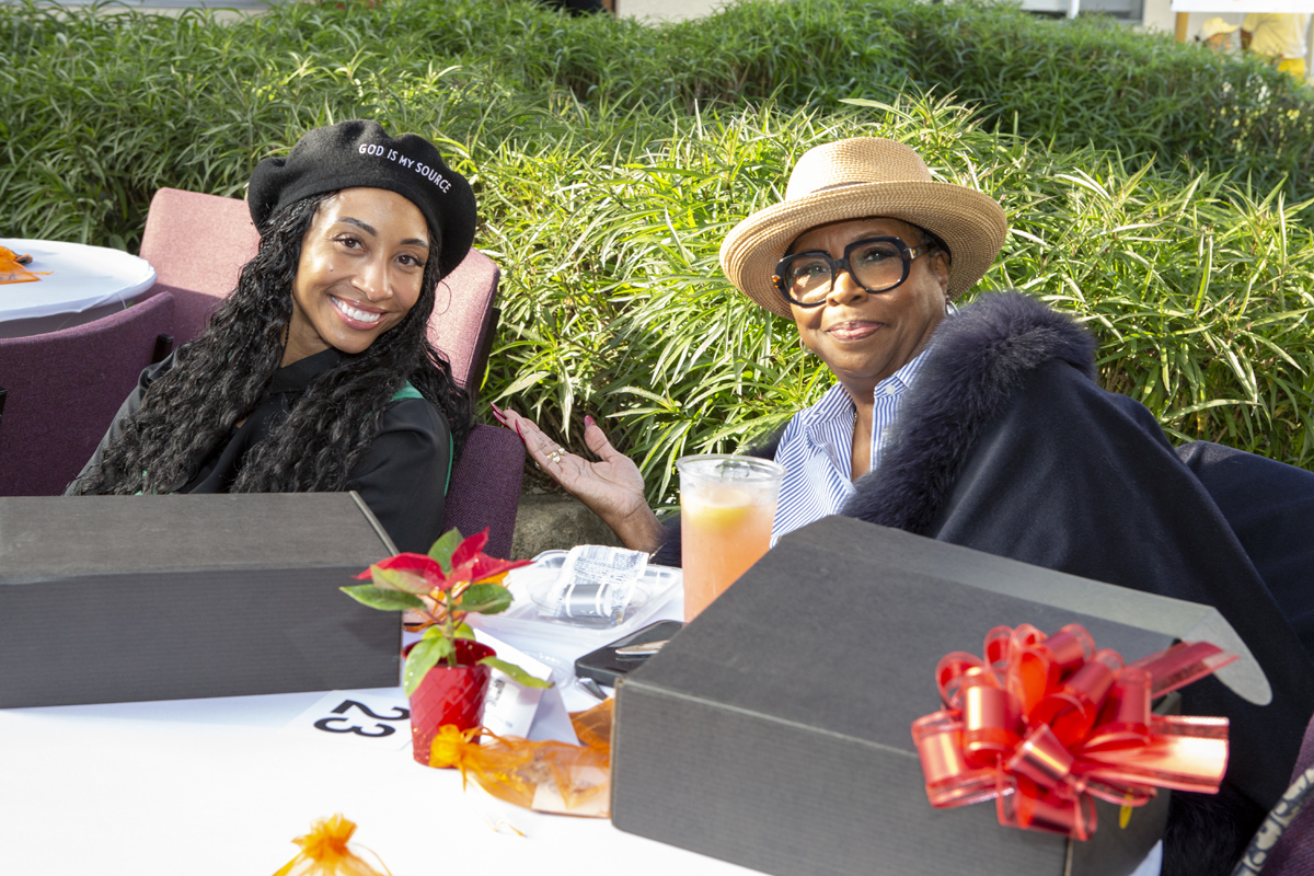Two women sitting at a table outdoors, smiling, with a gift box with a red bow, a potted plant, and drinks, surrounded by greenery.