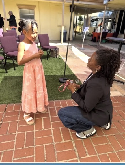 A young girl in a pink dress stands outdoors, talking to a woman kneeling on brick pavement, holding a small object.