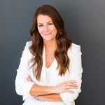 Woman with long brown hair smiling, arms crossed, wearing a white blazer, standing against a dark background.