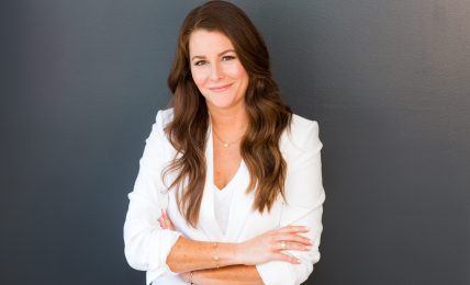 Woman with long brown hair smiling, arms crossed, wearing a white blazer, standing against a dark background.