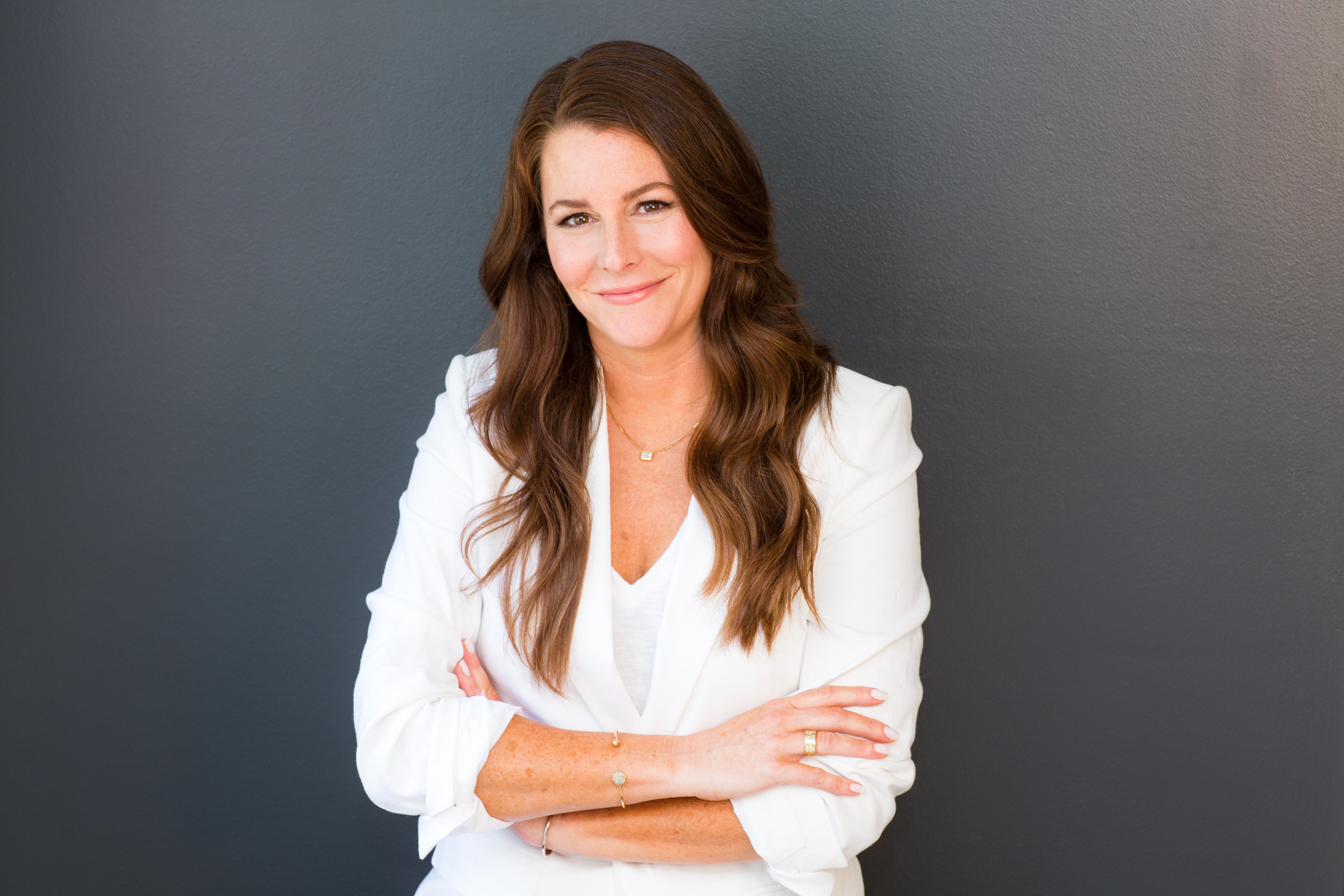 Woman with long brown hair smiling, arms crossed, wearing a white blazer, standing against a dark background.