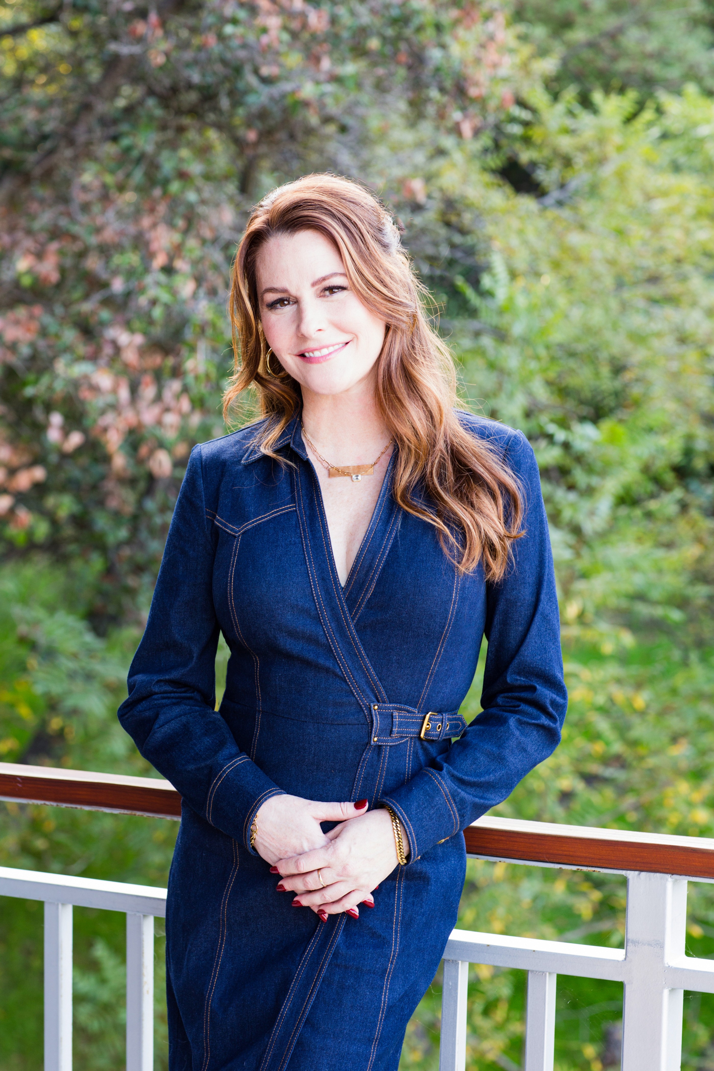 Young woman with long wavy hair smiling outdoors, wearing a dark blue dress, standing by a white railing with green trees behind her.
