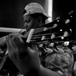 Person playing guitar, wearing a cap backwards, in a dark setting, focused on the instrument.