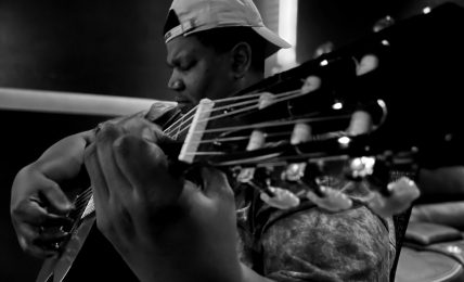 Person playing guitar, wearing a cap backwards, in a dark setting, focused on the instrument.