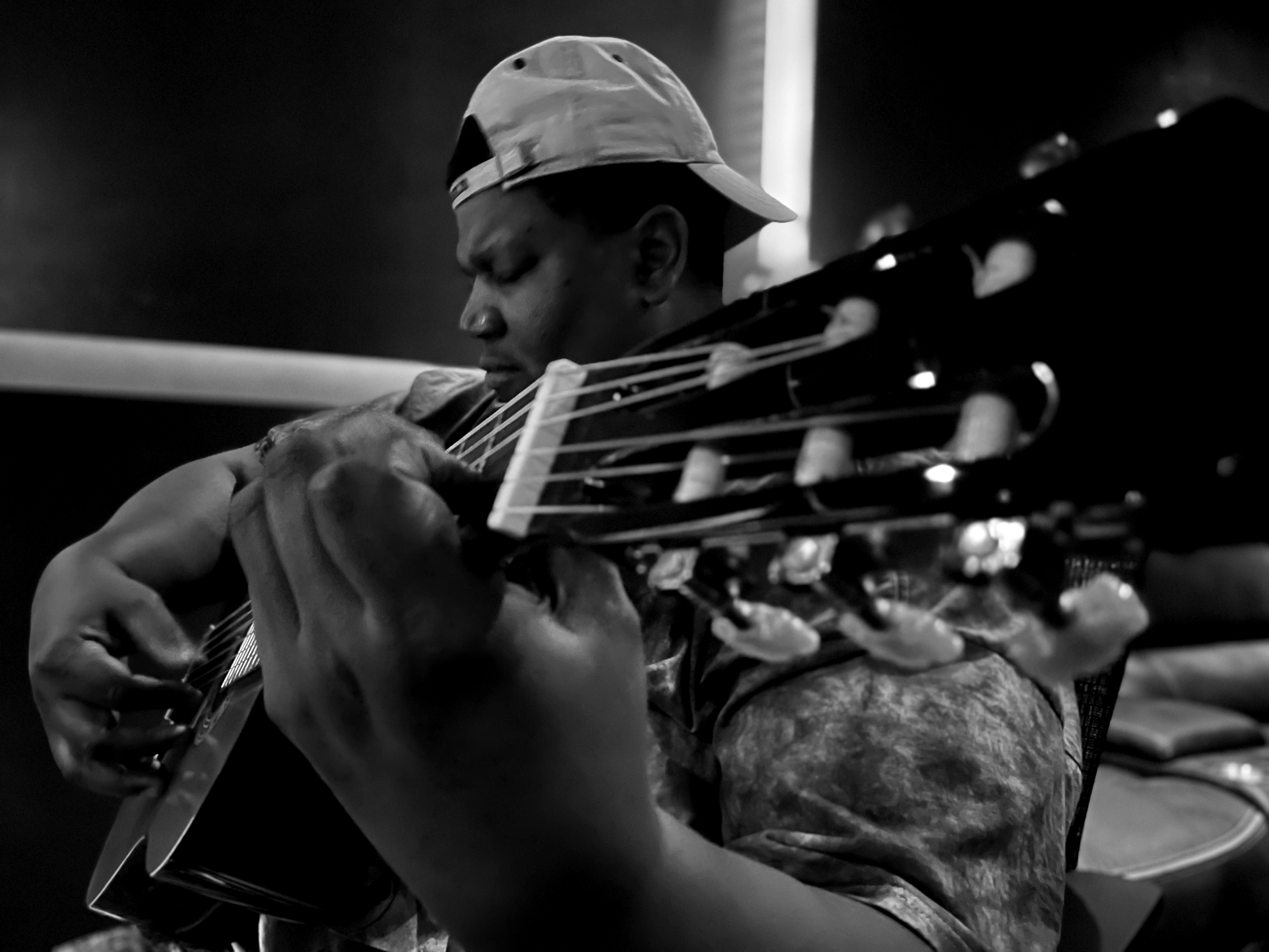 Person playing guitar, wearing a cap backwards, in a dark setting, focused on the instrument.