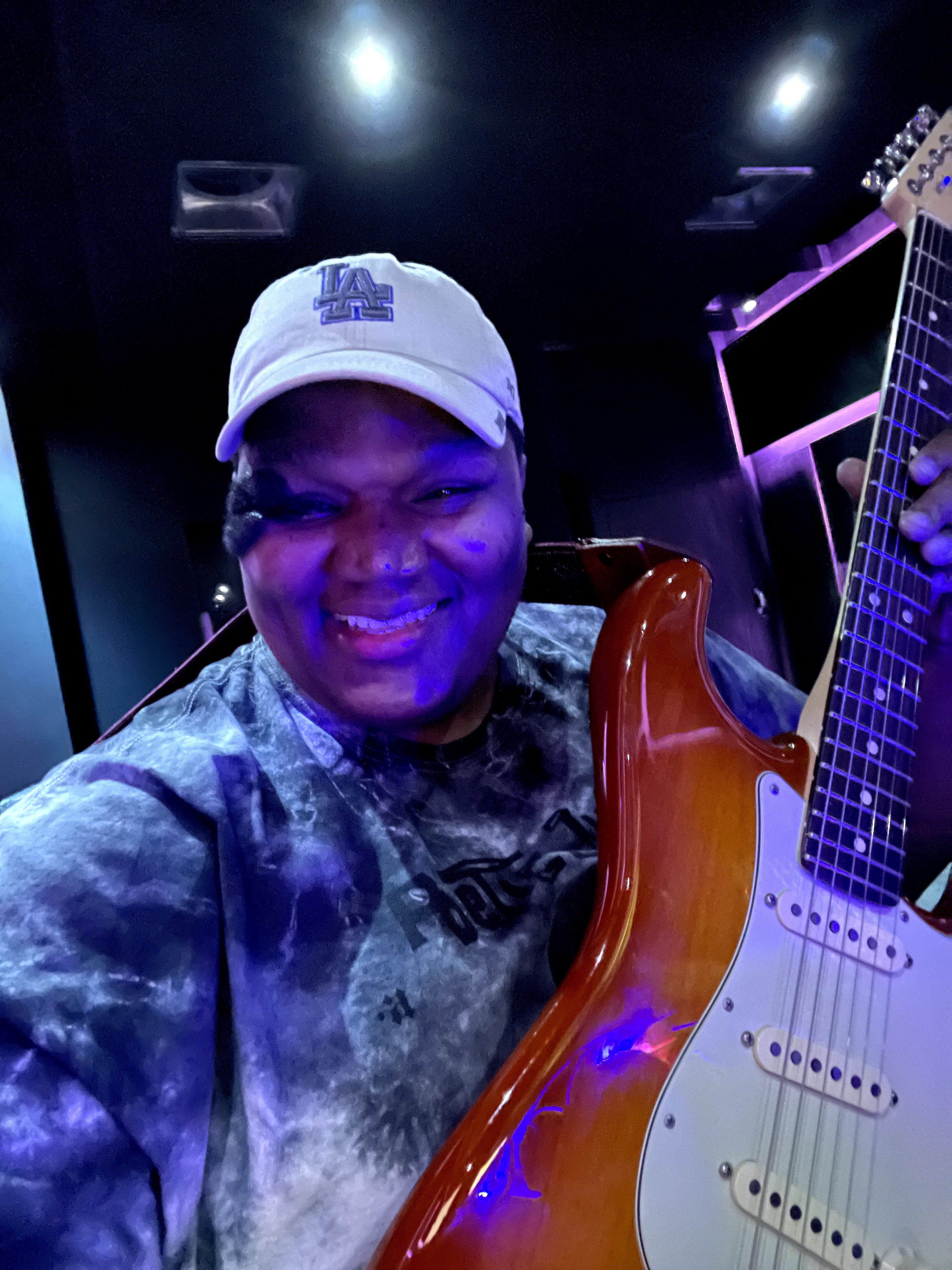 Person smiling, wearing a white cap, holding an electric guitar, in a dark room with purple lighting.
