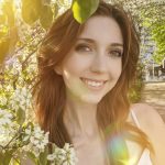 Young woman smiling outdoors with blooming flowers and sunlight, surrounded by green leaves and trees.