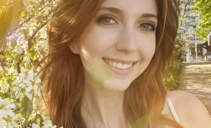 Young woman smiling outdoors with blooming flowers and sunlight, surrounded by green leaves and trees.
