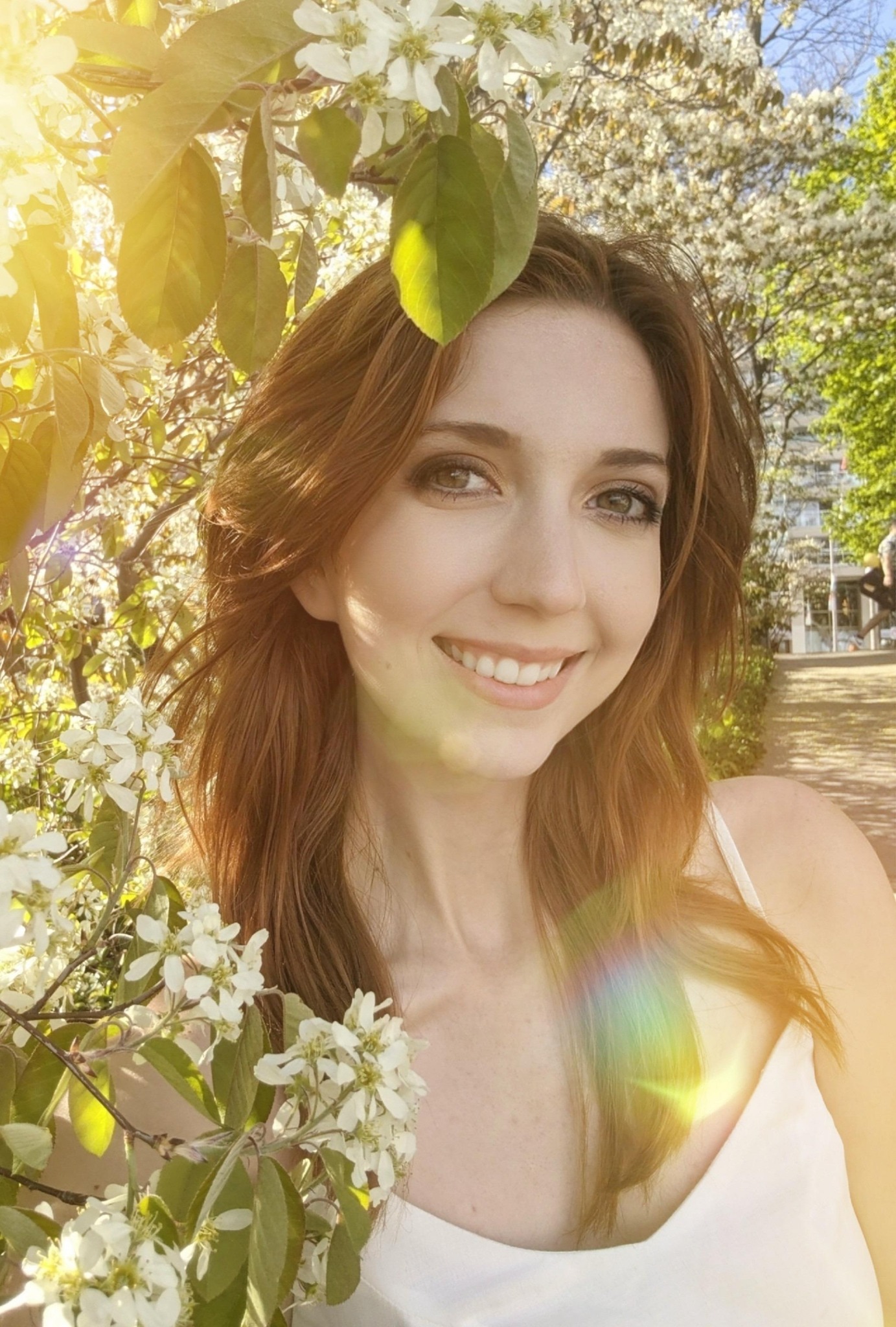 Young woman smiling outdoors with blooming flowers and sunlight, surrounded by green leaves and trees.