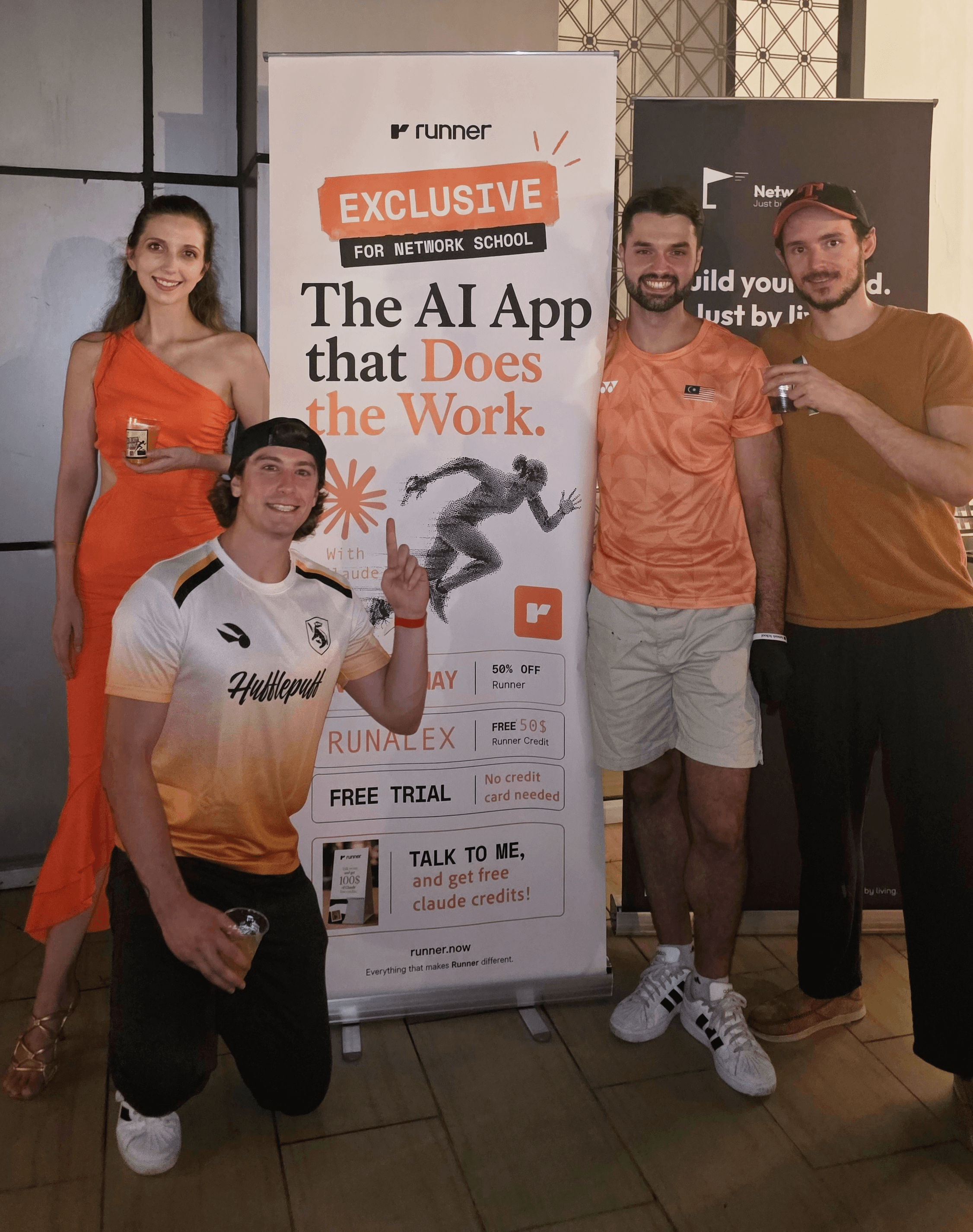 Four people standing next to a promotional banner, smiling and posing indoors. One person kneeling in front, three standing behind.
