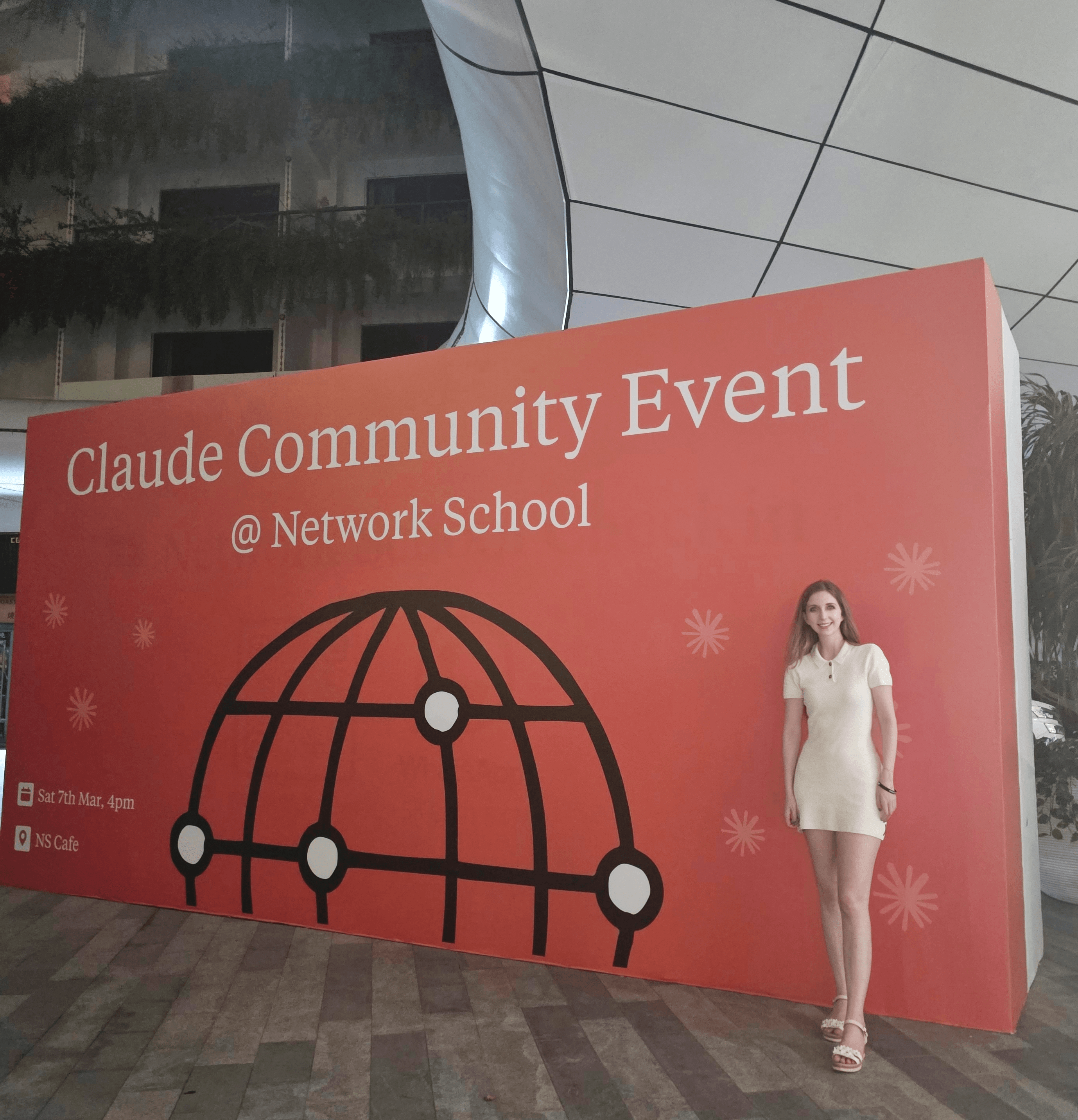 Large red sign for Claude Community Event at Network School with a woman standing beside it, outdoors at night.
