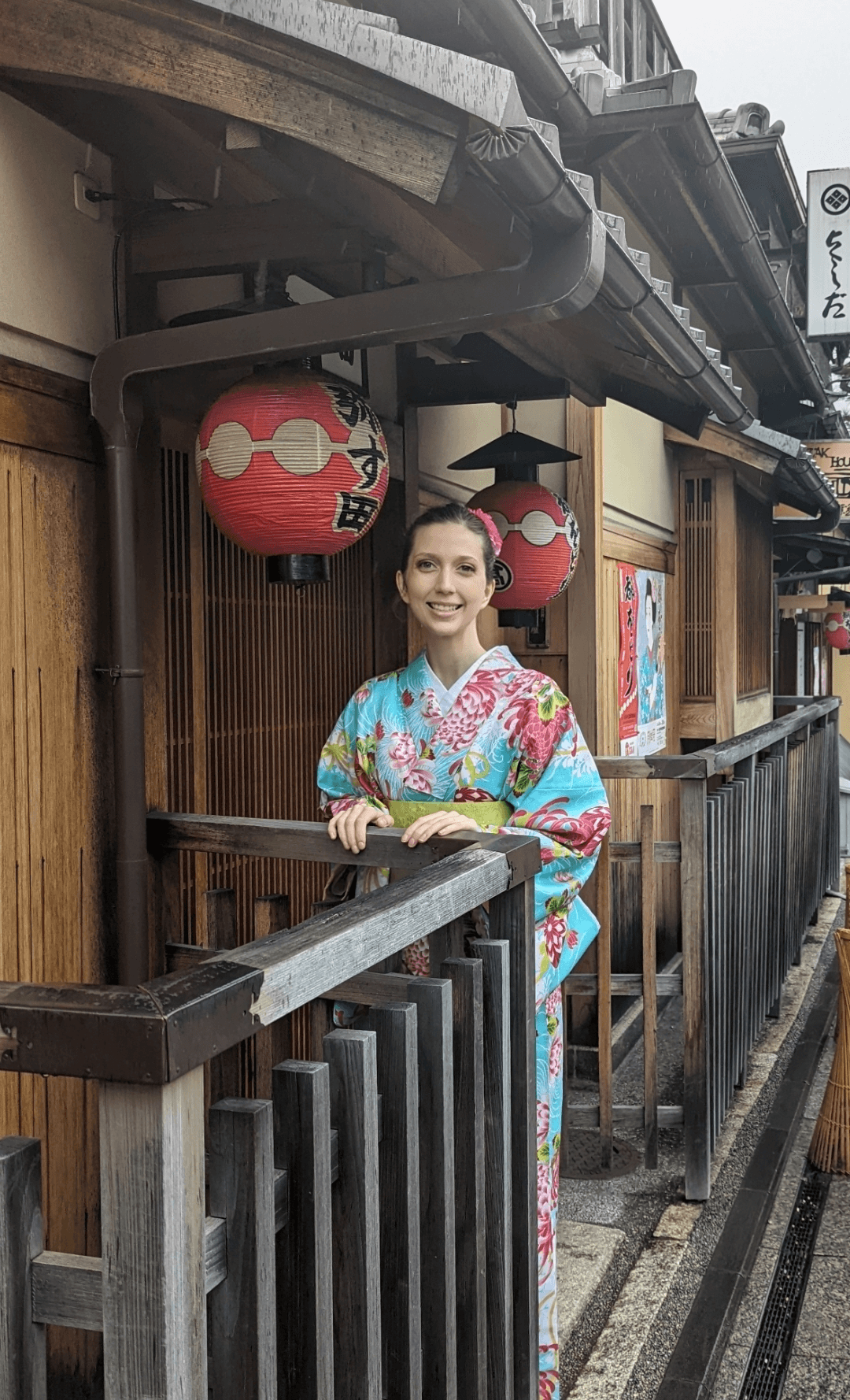 Woman in colorful kimono standing on wooden balcony with red lanterns behind her, traditional building in background.