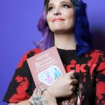 Woman with colorful hair holding a book, smiling, against a purple background.