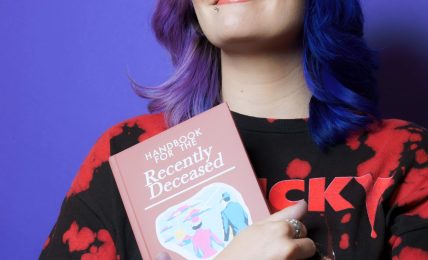 Woman with colorful hair holding a book, smiling, against a purple background.