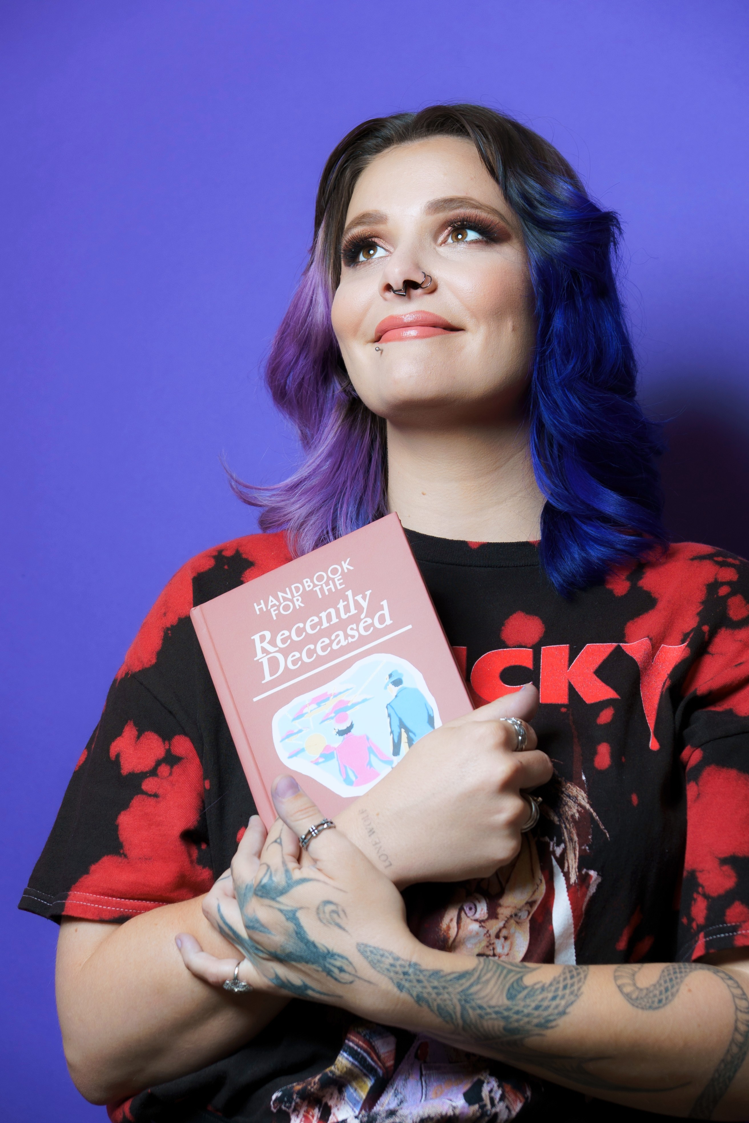 Woman with colorful hair holding a book, smiling, against a purple background.