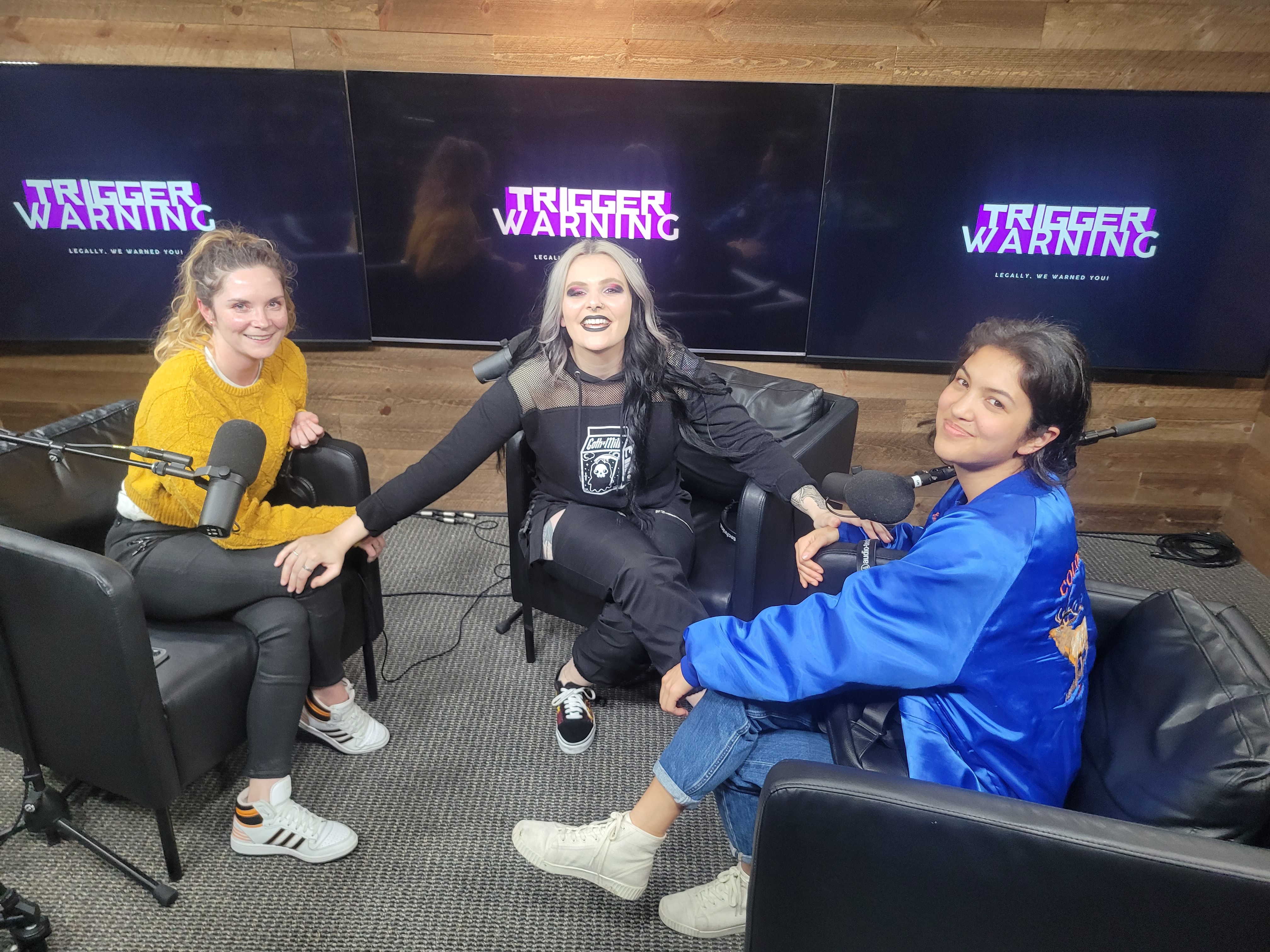 Three people sitting in a studio with multiple screens displaying 'TRIGGER WARNING' behind them, smiling and holding hands.