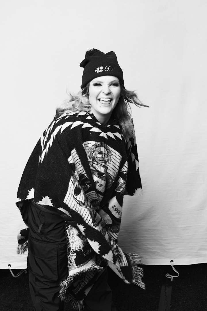 Woman smiling, wearing a beanie and patterned poncho, standing against a plain background.