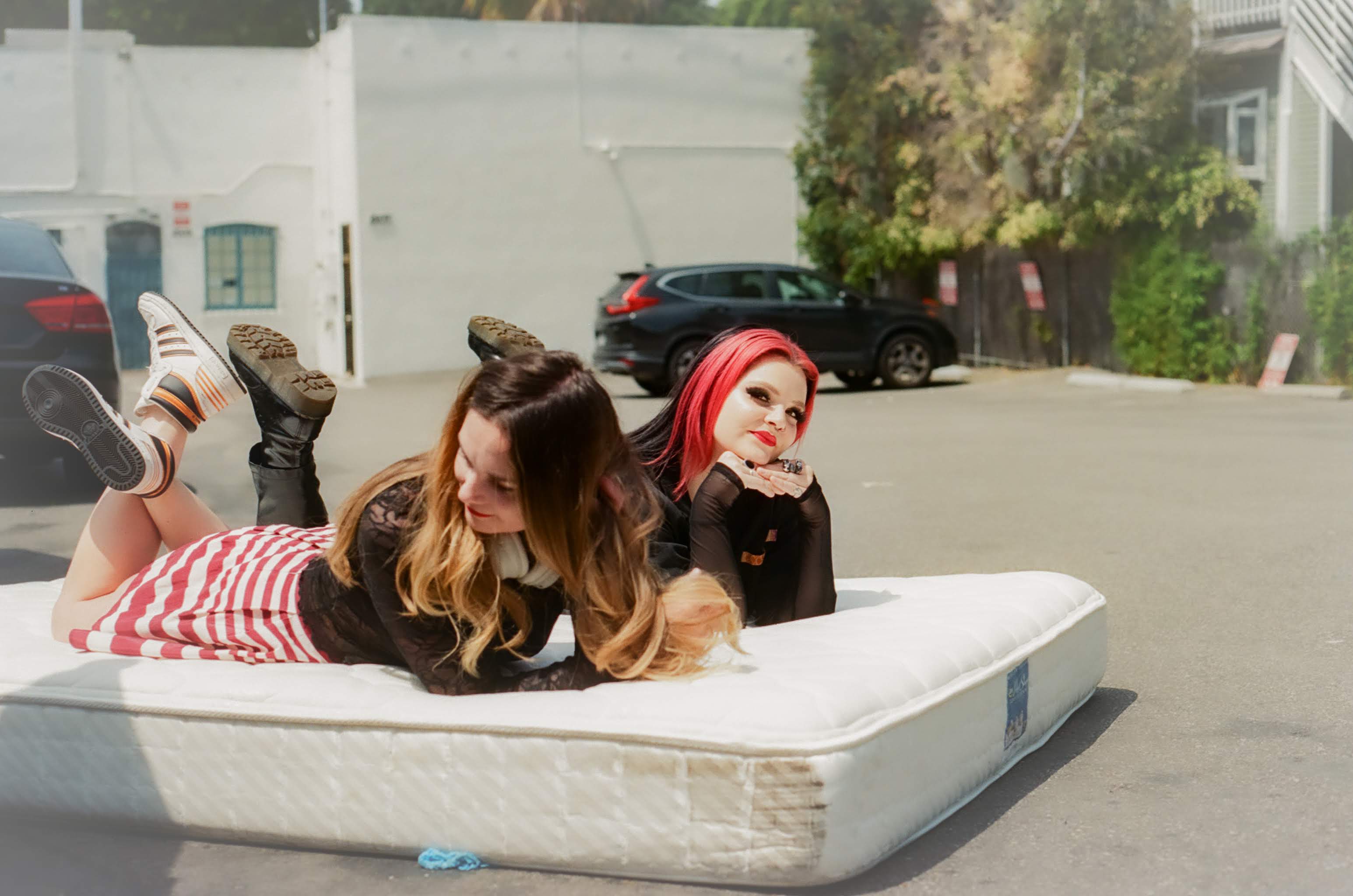 Two women lie on a mattress outdoors, one with long brown hair and the other with bright red hair, in a parking lot.