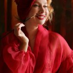 Woman with red hair and lipstick, wearing a red silk robe, smiling and holding a cigarette holder.