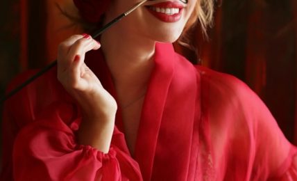Woman with red hair and lipstick, wearing a red silk robe, smiling and holding a cigarette holder.