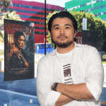 Man with black hair and beard wearing a white shirt, standing outdoors with colorful buildings in background.