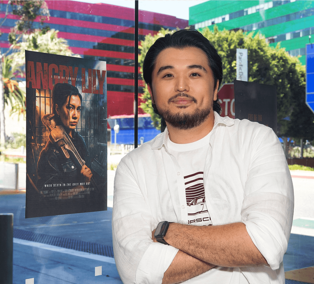 Man with black hair and beard wearing a white shirt, standing outdoors with colorful buildings in background.