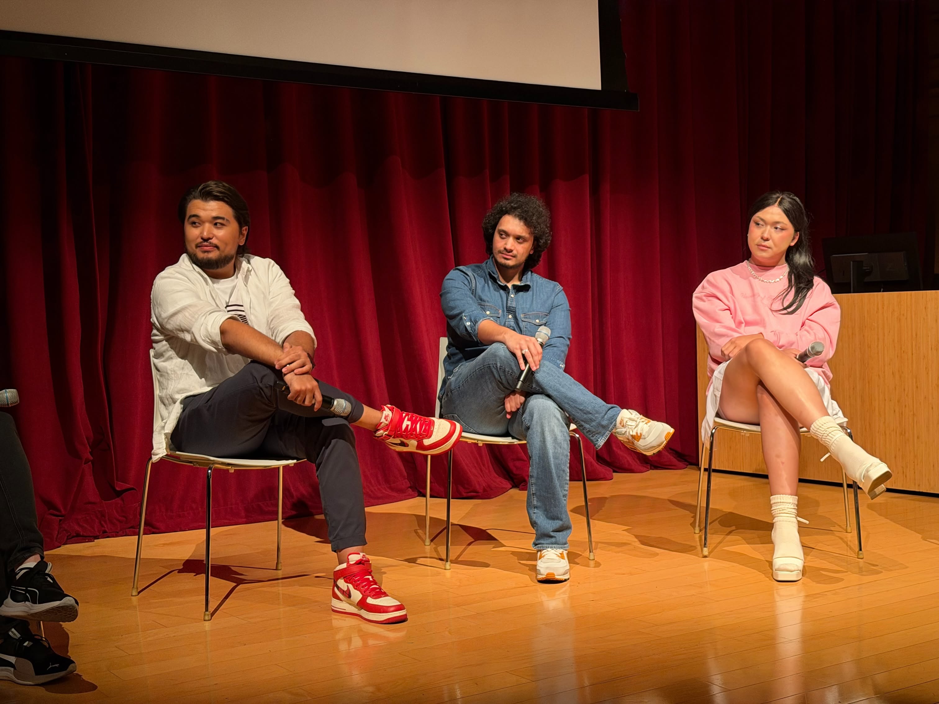 Three people sitting on chairs on a stage with a red curtain background, engaged in a discussion.