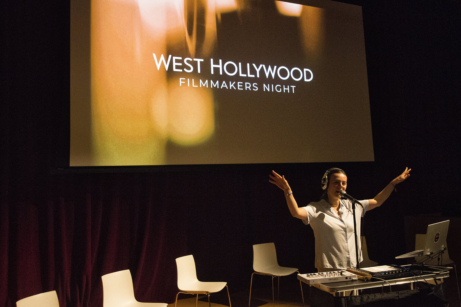 Woman speaking on stage with arms raised at West Hollywood Filmmakers Night event, large screen behind her, chairs on stage.