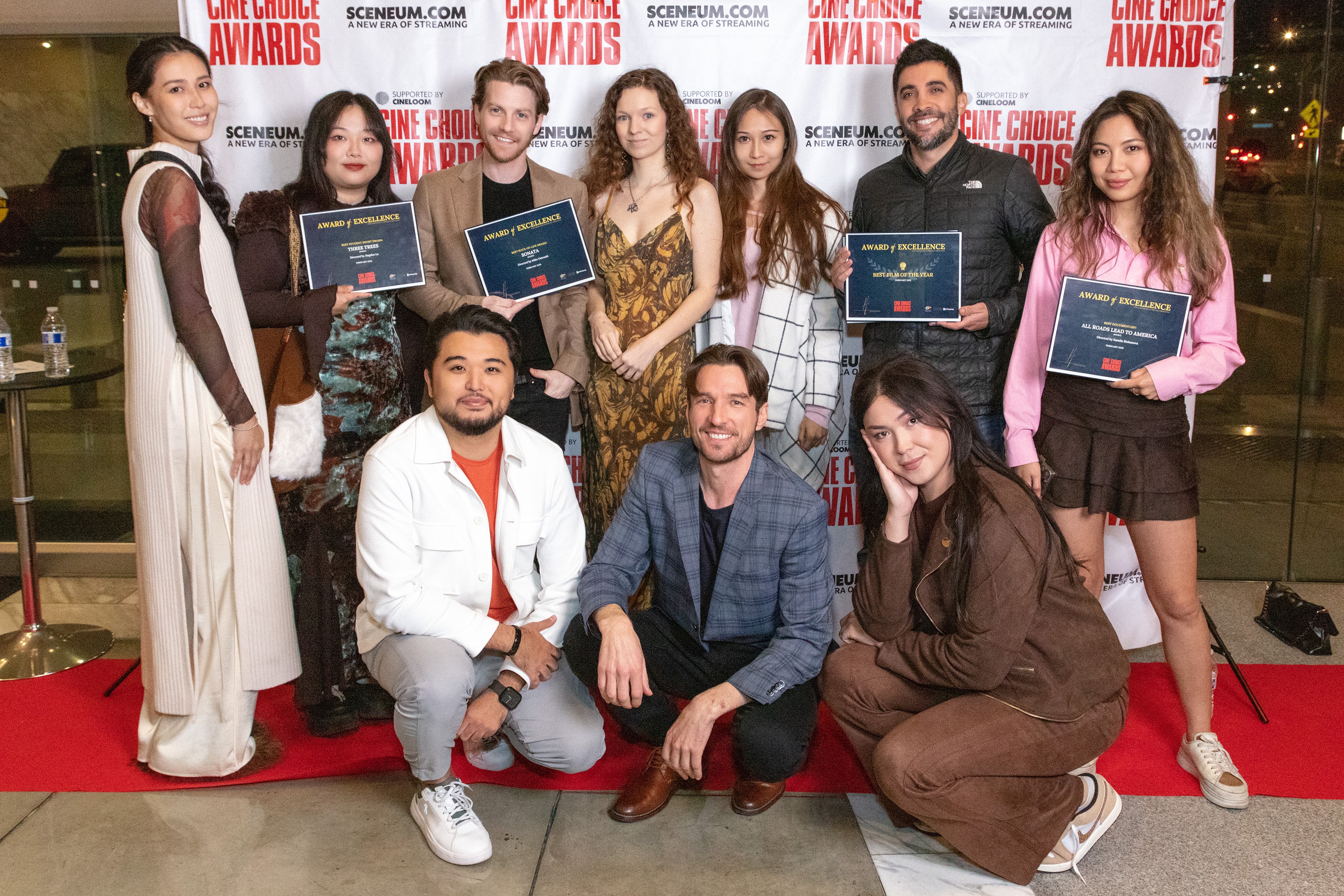 Group of nine people, some holding awards, standing and sitting on a red carpet at an event, with a backdrop of logos.