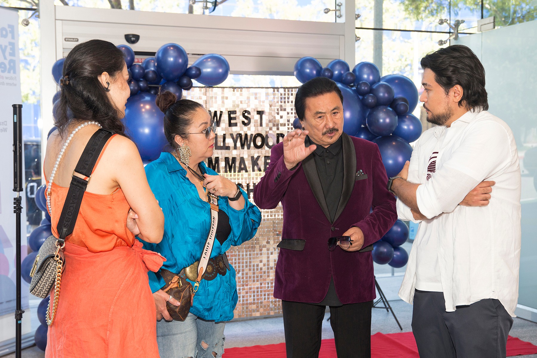 Four people engaged in conversation indoors, with blue balloons and a sign in the background, sunlight streaming through windows.